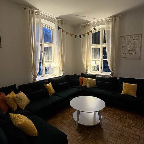 Gemütliches Wohnzimmer mit einem dunklen Ecksofa, gelben und orangefarbenen Kissen, einem weißen runden Couchtisch, Lampen, Vorhängen und einer Girlande aus Faden. In der Nähe der Eckfenster hängt ein Wandbehang mit Schriftzug.