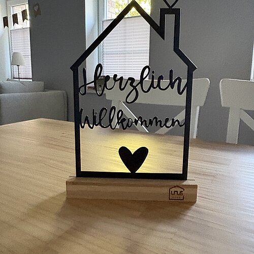 Ein Metallschild in Form eines Hauses mit der Aufschrift „Herzlich Willkommen“ und Herzmotiven auf einem Holzsockel, das auf einem Tisch in einem gemütlichen, sonnendurchfluteten Raum steht.