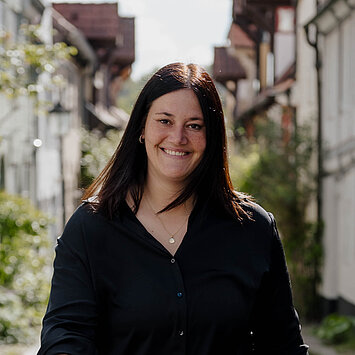 Eine Frau mit langen dunklen Haaren und einer Tätowierung am linken Arm geht lächelnd in einem schwarzen T-Shirt und Jeans auf einer Kopfsteinpflasterstraße zwischen historischen Gebäuden entlang.
