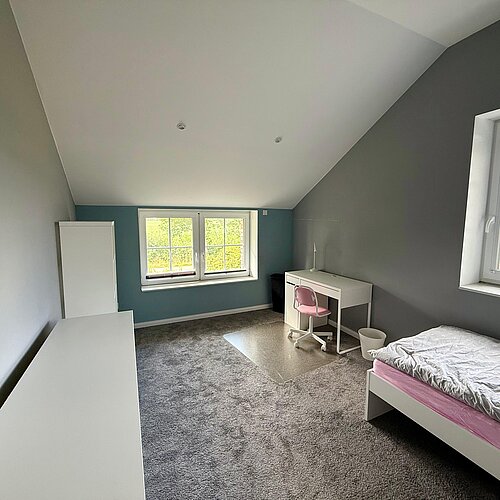 Ein minimalistisch eingerichtetes Schlafzimmer mit einem Einzelbett, einem Schreibtisch, einem rosa Stuhl, einem weißen Kleiderschrank und großen Fenstern, durch die Tageslicht hereinströmt. Die Wände sind blau und grau gestrichen, und auf dem Boden liegt Teppichboden.