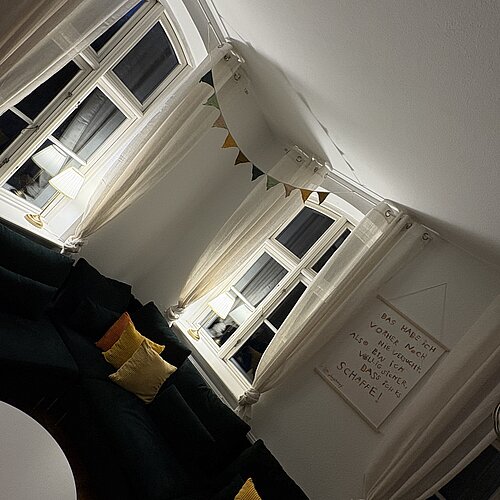 Ein gemütliches Wohnzimmer mit weißen Wänden, großen Fenstern, grünen Sofas mit gelben Kissen, Lichterketten, einer dreieckigen Wimpelkette und einem Wandbehang mit Schriftzug, alles sanft von Lampen beleuchtet.