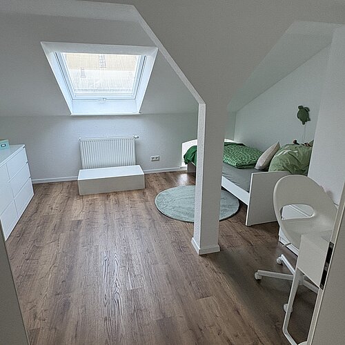 Ein helles Schlafzimmer im Dachgeschoss mit Holzboden, einem Dachfenster über einem Heizkörper, einem Bett in Grüntönen, einer weißen Kommode, einem weißen Stuhl und einem kleinen runden grünen Teppich.