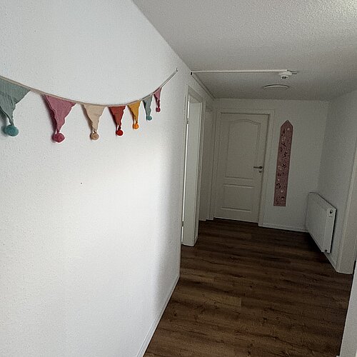 Ein Flur mit Holzboden, weißen Wänden, einer bunten dreieckigen Girlande auf der linken Seite, einer geschlossenen weißen Tür am Ende und einem Heizkörper an der rechten Wand.
