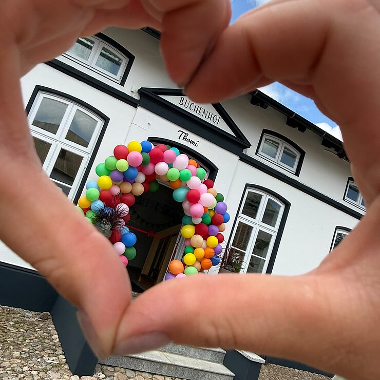 Hände bilden ein Herz und umrahmen ein weißes Gebäude mit einem gewölbten Eingang, der mit bunten Luftballons geschmückt ist. Der Boden besteht aus Kopfsteinpflaster, und der Himmel ist teilweise bewölkt.