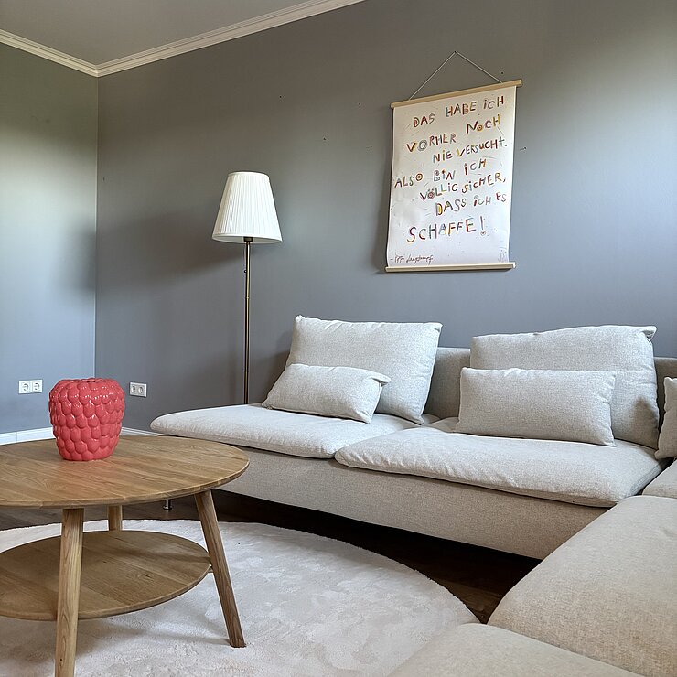 Ein modernes Wohnzimmer mit einem beigen Ecksofa, einem Couchtisch aus Holz mit einer roten Vase, einem hellen Teppich und einer Stehlampe. An der grauen Wand über dem Sofa hängt ein handgeschriebenes Schild auf Deutsch.