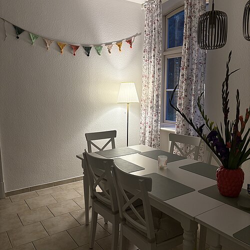 Ein gemütliches Esszimmer mit einem weißen Tisch und Stühlen, geblümten Vorhängen, einer Stehlampe, Hängelampen, einer bunten Wimpelkette und einer Vase mit leuchtend bunten Blumen auf dem Tisch.