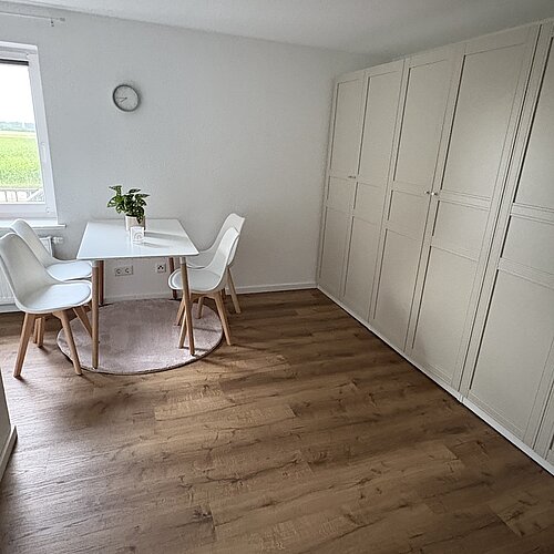 Ein minimalistisch eingerichtetes Zimmer mit Holzboden, einem weißen Kleiderschrank, zwei weißen Stühlen, einem kleinen weißen Tisch mit einer Pflanze, einer Wanduhr und einem Fenster, durch das Tageslicht hereinfällt.
