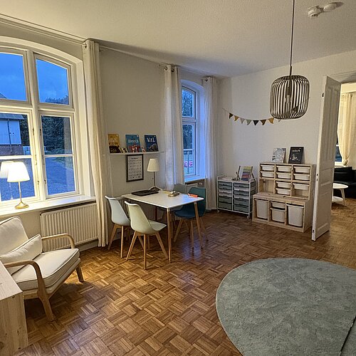 Ein gemütliches Zimmer mit Holzböden, einem runden Teppich, einem kleinen Tisch mit Stühlen, Büchern in den Regalen, einem bequemen Sessel, Aufbewahrungskörben und großen Fenstern mit weißen Vorhängen, durch die das Abendlicht hereinfällt.